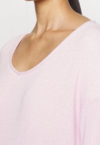 Vaaleanpunainen, tekstuurinen neuletakki, jossa on leveä avonainen kaula-aukko. Pehmeä neulottu kangas on löysästi istuva, mikä tekee siitä täydellisen kerrospukeutumiseen.