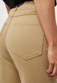 Bež hlače visokog struka s elegantnim dizajnom, koje imaju središnji šav, vezane džepove i teksturiranu pamučnu tkaninu.