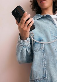 Person i denimjakke, der holder en sort smartphone med ring og kæde vedhæftet, imod en ensartet lys baggrund.