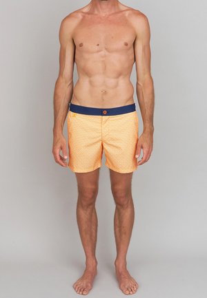 Shorts de bain oranges avec un motif géométrique, présentant une taille en bleu foncé et un bouton à l'avant. Le matériau semble léger.