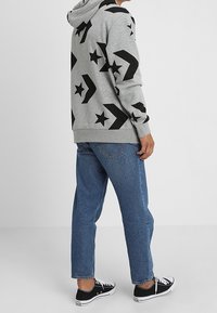 Gray hoodie with black star and arrow patterns, paired with blue denim jeans and black sneakers. The hoodie features a drawstring hood.