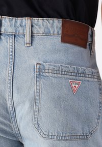Šviesiai mėlyni džinsai šortai su odiniu Guess prekės ženklo lipduku ir raudonu trikampio logotipu ant užpakalinio kišenės. Matoma detalizuota siuvimo apdaila.