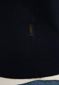 Närbild av marinblått stickat tyg med en liten svart etikett märkt "CHASIN'" i orangea bokstäver nära sömmen.