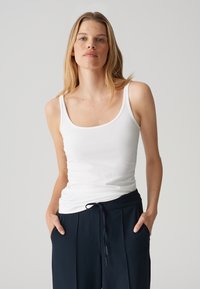 Top bianco senza maniche con spalline sottili, presenta un collo a barchetta. Tessuto morbido e liscio; indossato con pantaloni blu navy dotati di vita regolabile con cordino.