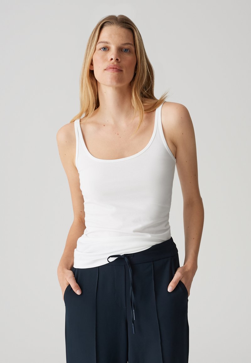 Top bianco senza maniche con spalline sottili, presenta un collo a barchetta. Tessuto morbido e liscio; indossato con pantaloni blu navy dotati di vita regolabile con cordino.