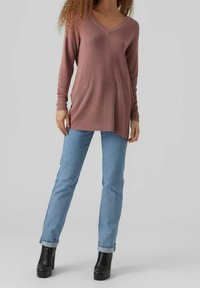 Femme portant un pull tunique mauve à manches longues avec un col en V, un jean bleu clair retroussé et des bottines à plateformes noires, debout sur un fond gris.