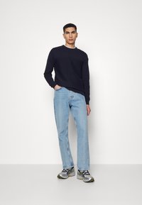 Marine gebreide trui, lichtblauwe rechte jeans en grijze sneakers met zwarte en gele accenten, staand tegen een effen witte achtergrond.