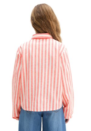 Osoba o długich brązowych włosach, ubrana w różowo-białą pionowo paskowaną koszulę z długim rękawem i niebieskie dżinsy, widziana od tyłu.