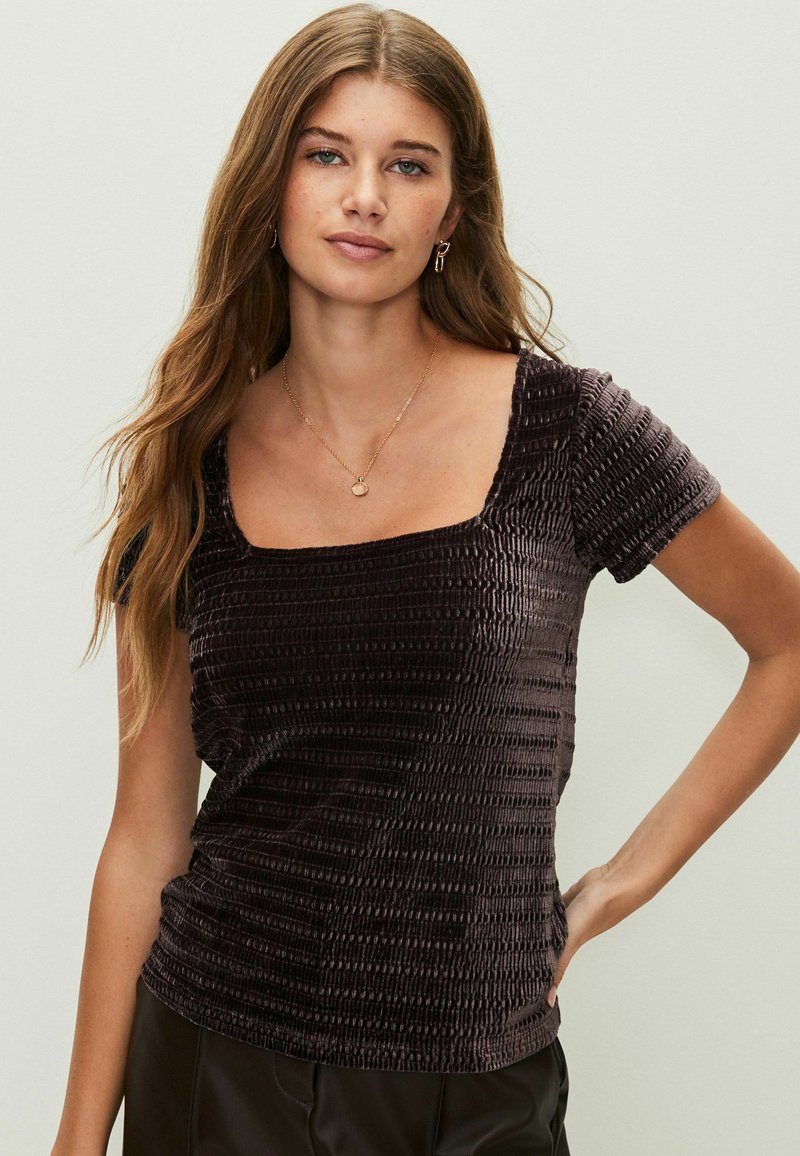 Sötét bársony blúz négyzet alakú nyakkivágással, rövid, ráncolt ujjakkal, texturált felülettel és fitted szabással, minimalista arany ékszerekkel párosítva.