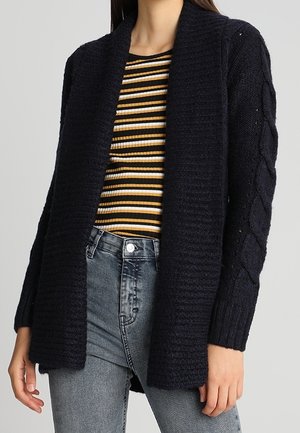 Frau trägt eine schwarze, offene Strickjacke mit Zopfmuster über einem gelb-weiß-schwarz gestreiften, gerippten Oberteil und ausgewaschenen, hoch taillierten grauen Jeans.