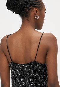 Fekete flitteres ruha vékony pántokkal, pikkelyszerű mintával és sima textúrával, részletes kidolgozással a bodzén.