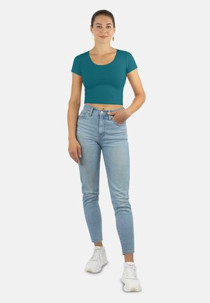 Teal gekürztes T-Shirt mit kurzen Ärmeln, kombiniert mit hochtaillierten hellblauen Jeans. Weiße Sportschuhe; Modell mit zusammengebundenem Haar.