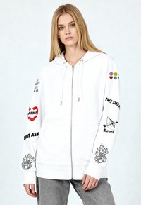 Felpa con cappuccio bianca con zip, caratterizzata da testo grafico e simboli in nero con accenti colorati, vestibilità comoda e tasca a marsupio.