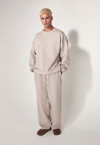 Sudadera oversize beige y pantalones a juego en tejido suave, con puños acanalados, cuello redondo y un sutil detalle de logo. Zapatos marrones tipo slip-on.
