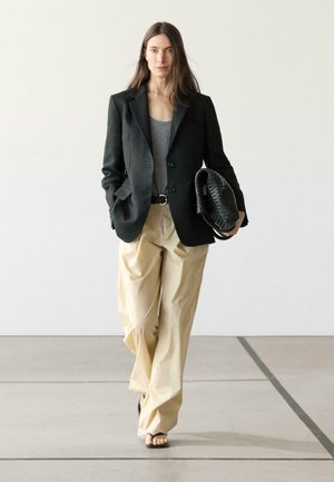 Frau geht drinnen und trägt einen schwarzen Blazer, ein graues Oberteil, beige weite Hosen und hält eine strukturierte schwarze Handtasche, die Hände teilweise in den Taschen.