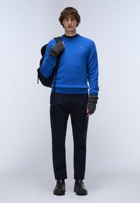 Mans blårandig tröja, stickad material, rundad halsringning, ribbad nederkant. Svart ryggsäck, grå fingerlösa handskar, marinblå byxor och svarta stövlar.