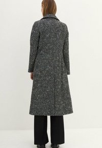 Cappotto grigio lungo con un motivo tessuto a trama, con rever a punta e maniche lunghe. Abbinato a pantaloni neri a gamba larga e stivali.