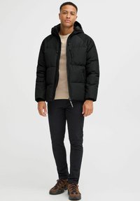 Sort pufferjakke med hætte, med lynlås og to sidelommer, iført over en beige sweater og sorte bukser.
