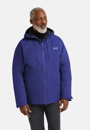 Mann mit grauem Bart, der eine blaue Jack Wolfskin Kapuzenjacke und schwarze Hose trägt und leicht seitlich schaut vor einem einfarbigen Hintergrund.