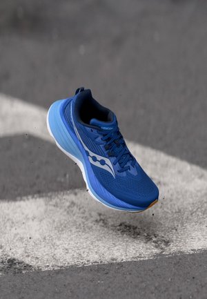 Blå Saucony løbesko i luften over en grå asfaltflade med en hvid malet linje, set fra siden og lidt ovenfra.