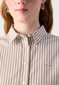 Una camicia button-up a righe con righe verticali marrone chiaro e bianche, colletto classico e un piccolo logo ricamato sul lato sinistro.