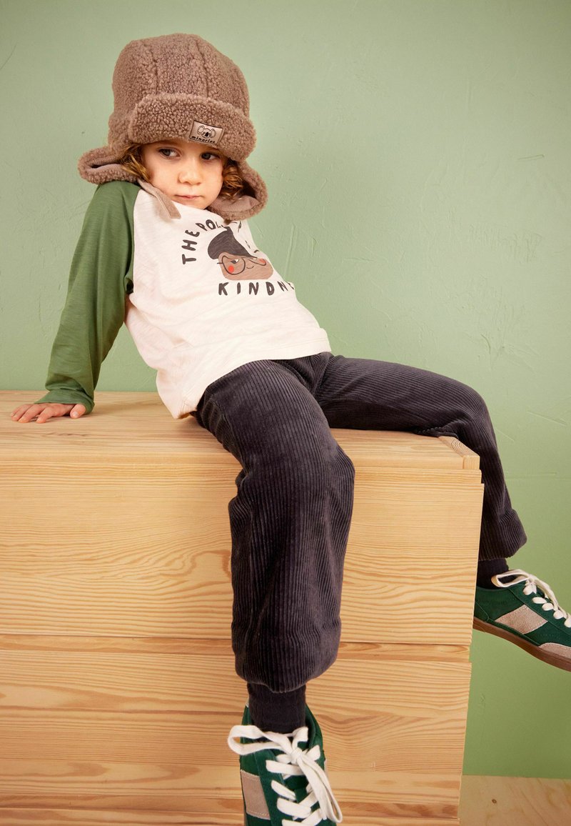 Lapsi istuu puulaatikolla, päällään ruskea fleece-hattu, vihreä pitkähihainen paita, tummansiniset samettihousut ja vihreät lenkkarit.