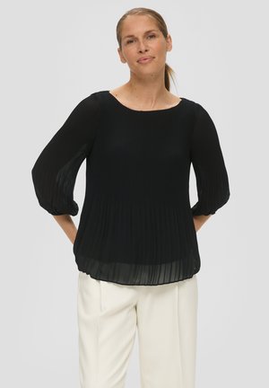 Zwarte geplooide blouse met driekwart mouwen, ronde halslijn en een vloeiende silhouette, gecombineerd met crèmekleurige broeken.