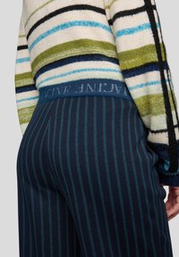 Gestreifter Strickpullover mit grünen, blauen und schwarzen Mustern; gerippte Bündchen und ein hoher Taillenbund mit kontrastierendem Logo an marineblauen Nadelstreifenhosen.