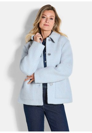 Frau mit langen blonden Haaren, die eine hellblaue, flauschige, geknöpfte Jacke über einer dunklen Jeans vor einem schlichten Hintergrund trägt.