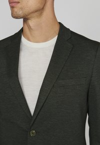 Dark green blazer made of textured fabric, featuring a one-button closure, a notch lapel, and a chest pocket over a white crew neck shirt.