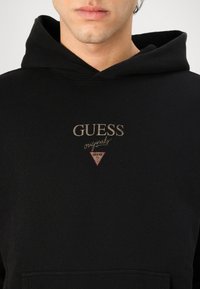 Čierna bavlnená mikina s kapucňou, obsahujúca klokanie vrecko, predný logo v zlatom písmene a červený trojuholníkový akcent pod názvom značky.