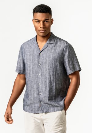 Mann trägt ein kurzärmliges, graues Hemd mit dünnen weißen vertikalen Streifen und hellfarbige Hose, steht vor einem schlichten Hintergrund.