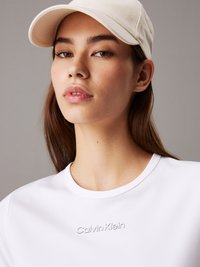 Bijela majica s teksturiranom tkaninom i utisnutim logom "Calvin Klein" na prsima, uparena s bež kapom.