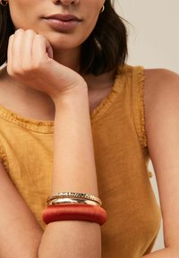 Guld- och orangearmband på en ljus hy-handled. I bakgrunden syns en texturerad, senapsgul topp med en fransad halsringning.