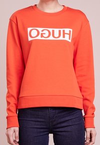 Pomarańczowy sweter z białym logo w formie prostokąta "HUGO". Okrągły dekolt, długie rękawy, dopasowany dół, gładka tekstura, luźny krój.