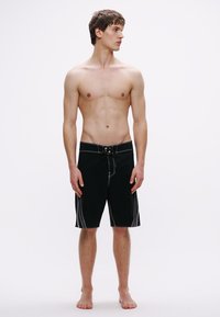 Μαύροι κοντοί αθλητικοί σορτς με λευκές ραφές, μέση με κορδόνι και πλαϊνές τσέπες· κατασκευασμένοι από ύφασμα κατάλληλο για δραστηριότητες στο νερό.