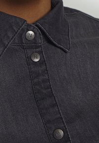 Dunkelblaue Jeanshemd mit strukturiertem Kragen, silbernen Knöpfen mit einem "N"-Logo und subtiler Textur im Stoff.