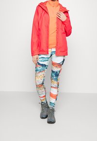 Veste imperméable rouge, haut intermédiaire orange, leggings à motifs multicolores et chaussures de randonnée grises. Présente des couleurs vives et des textures variées.