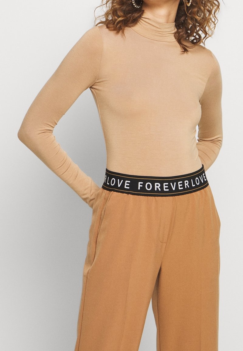 Maglione a maniche lunghe color tan con colletto alto abbinato a pantaloni beige larghi con una cintura nera con la scritta bianca "FOREVERLOVE." Tessuto liscio.