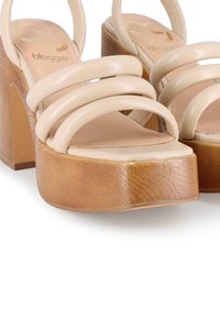 Sandalias de plataforma beige que presentan tres correas brillantes y un tacón de bloque de madera con un acabado texturizado. Forro interior suave y minimalismo en la marca.