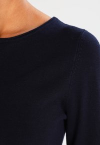 Tummansininen istuva yläosa pyöreällä pääntiellä, jossa on sileä neulottu pinta ja hienovaraiset saumakoristelut olkapäillä.