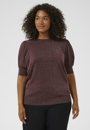 Woman with curly hair wearing a glittery plum-colored top with puffed short sleeves and black pants, looking slightly to the side.