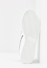 Bílé tenisky s texturovanou gumovou podrážkou s cikcak vzory. Na boku je černý pruh a detail loga na patě.