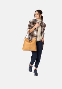 Ruskea nahkainen hobo-laukku, jossa on pehmeä muotoilu ja ommeltu yksityiskohta, käytetään olalla, yhdistettynä ruudulliseen paitaan ja rentoon asusteeseen.