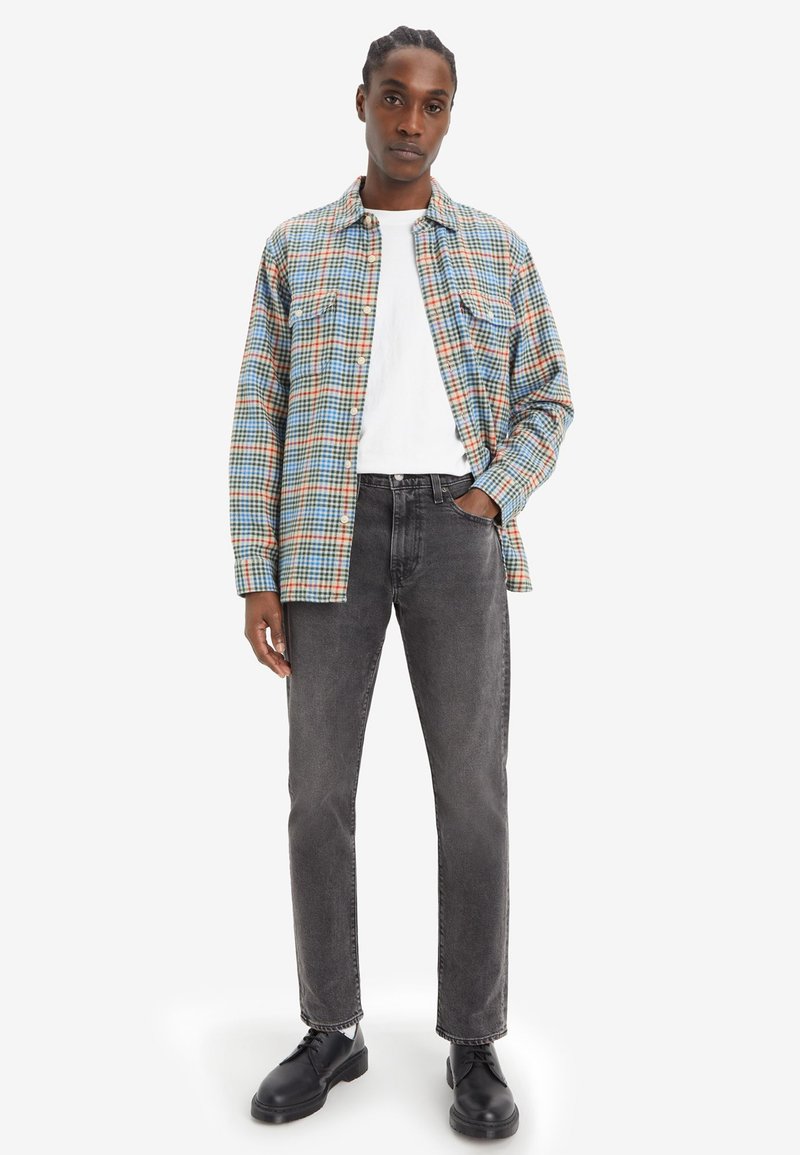 Chemise flanelle à carreaux bleus, orange et crème, superposée sur un t-shirt blanc. Un jean gris et des chaussures noires complètent la tenue.