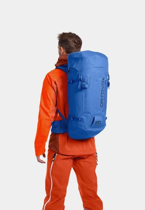 Blaue Rucksack mit Rolltop-Design, robustem Stoff, verstellbaren Trägern und einem oberen Griff. Logo auf der Seite sichtbar.getragen von einer Person in einer orangefarbenen Jacke.