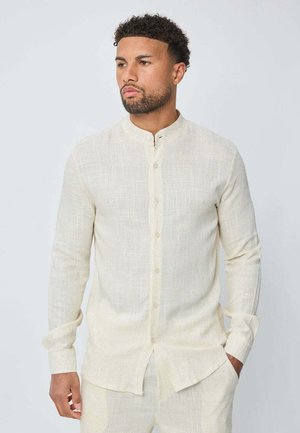 Chemise à manches longues de couleur crème en tissu texturé, avec un col montant et un devant à boutons, et un ourlet légèrement arrondi.