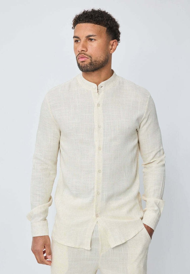 Chemise à manches longues de couleur crème en tissu texturé, avec un col montant et un devant à boutons, et un ourlet légèrement arrondi.