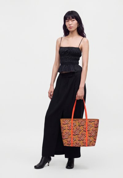 Femme portant un haut noir et un pantalon à jambes larges, tenant un sac cabas marron à motifs avec des anses orange, debout devant un fond clair uni.