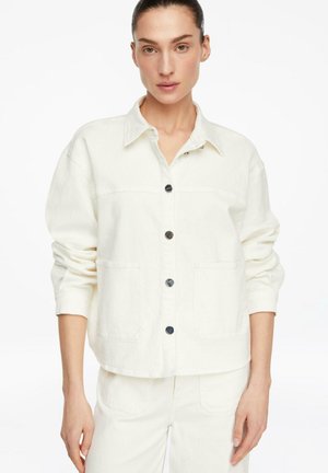 Kobieta z włosami związanymi do tyłu, ubrana w kremową kurtkę na guziki z dużymi przednimi kieszeniami oraz pasujące spodnie, na białym tle.